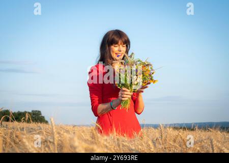 Hübsche Frau mit einem Blumenstrauß in der Hand steht erfreut in einem reifen Getreidefeld. Junge und attraktive Frau mit einem roten Kleid in einem reifen Getreidefeld genießt den Sommer. Region Weser-Ems Niedersachsen Deutschland *** hübsche Frau mit einem Blumenstrauß in der Hand glücklich auf einem Reifen Maisfeld stehend Junge und attraktive Frau mit rotem Kleid in einem Reifen Maisfeld genießt die Sommerregion Weser Ems Niedersachsen Deutschland Stockfoto