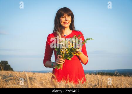 Hübsche Frau mit einem Blumenstrauß in der Hand steht erfreut in einem reifen Getreidefeld. Junge und attraktive Frau mit einem roten Kleid in einem reifen Getreidefeld genießt den Sommer. Region Weser-Ems Niedersachsen Deutschland *** hübsche Frau mit einem Blumenstrauß in der Hand glücklich auf einem Reifen Maisfeld stehend Junge und attraktive Frau mit rotem Kleid in einem Reifen Maisfeld genießt die Sommerregion Weser Ems Niedersachsen Deutschland Stockfoto