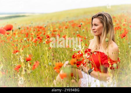 Blonde junge Frau, die Blumen auf einem Feld mit rotem Mohn pflückt. Stockfoto