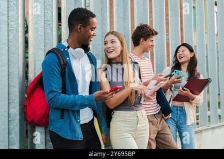 Gruppe glücklicher junger Studenten, die sich auf dem Universitätscampus unterhalten, lachen und Ideen austauschen, während sie Smartphones und Notebooks verwenden. Klassenkameraden, die Spaß mit einem Handy haben. Teenager mit Mobiltelefonen. Hochwertige Fotos Stockfoto