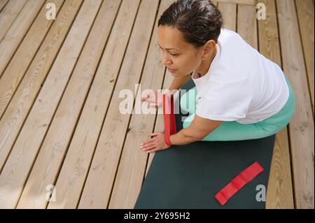 Eine Frau in einem weißen Hemd und einer grünen Hose übt Yoga auf einem Holzdeck mit einem roten Widerstandsband, wobei sie sich auf Fitness und Achtsamkeit konzentriert. Stockfoto