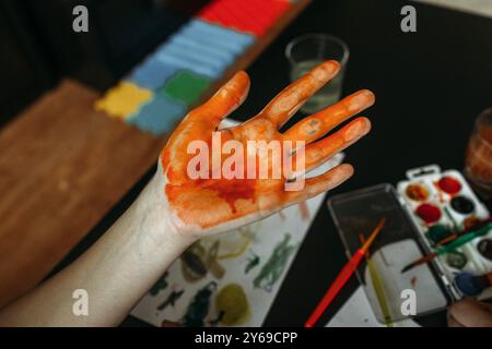 Eine Hand, die mit hellorangefarbener Farbe bedeckt ist, liegt auf einem schwarzen Tisch. Um sie herum gibt es verschiedene Kunstgegenstände, die einen freudigen Moment der Kreativität in einer gemütlichen Stockfoto