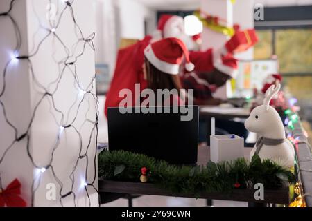 Nahaufnahme von Weihnachtslichtergirlanden, die die Hallen des Firmenbüros bedecken, mit Mitarbeitern, die Geschenke vom Weihnachtsmann in verschwommenem Hintergrund erhalten. Weihnachtsschmuck in festlich dekoriertem Arbeitsbereich Stockfoto