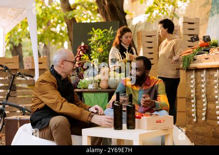 Schwarzer Mann sitzt am Tisch mit älteren Verkäufern und trinkt Biowein auf dem Bauernmarkt. Multirassische Menschen, die Spaß beim Essen ernten Festival haben, männliche Kunden und Bauern, die hausgemachte Getränke trinken. Stockfoto