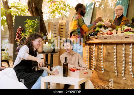 Glückliche kaukasische Frauen sitzen und trinken hausgemachten Wein auf dem lokalen Bauernmarkt. Weibliche Verkäuferin und Konsumentin verkostet frisches natürliches Bio-Produkt auf der Erntefarm Messe. Stockfoto