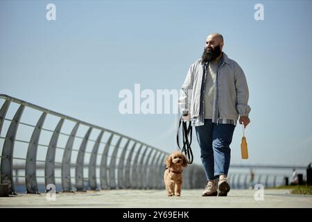 Ein Mann, der einen kleinen braunen Hund entlang eines Weges führt, der von geschwungenen Geländern vor klarem blauem Himmel begrenzt wird und die Tasche in der Hand hält Stockfoto