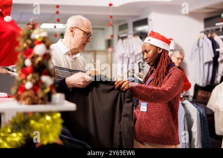 Der Mitarbeiter des Einzelhandels hilft dem alten Kunden bei der richtigen Kleidung und empfiehlt ermäßigte Artikel aus den Regalen. Ladenangestellte, die Kleidung für Senioren auswählte, weihnachtseinkaufsreise. Stockfoto