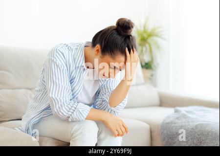 Stress, Kopfschmerzen, Überlastung, Depression. Frustrierte weißbrünette Frau sitzt nach einem harten Arbeitstag auf einem Sofa im Wohnzimmer, leidet unter Kopfschmerzen oder hat persönliche Probleme, fühlt sich einsam, braucht Ruhe Stockfoto