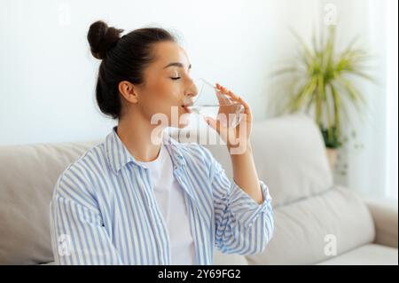 Glückliche positive arabische oder indische Brünette in legeren Kleidern trinkt am Morgen eine Tasse sauberes Wasser, während sie auf einer Couch im Wohnzimmer sitzt. Gesundes Lebensstilkonzept Stockfoto