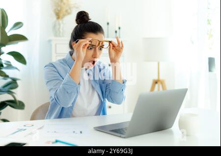 Überraschte, schockierte Frau arabischer oder indischer Nationalität mit Brille, Büroangestellte, schaut in verwirrter Hebebrille auf den Laptop-Bildschirm, ist verwirrt über die Informationen, die er sah Stockfoto