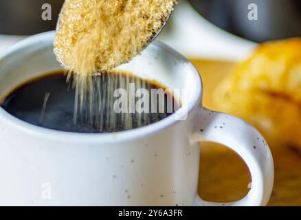 Demerara-Zucker, der aus einem silbernen Teelöffel in eine kleine Tasse heißen Espresso-Kaffee fällt, in einer Wohnküche, Worcestershire, Großbritannien. Stockfoto