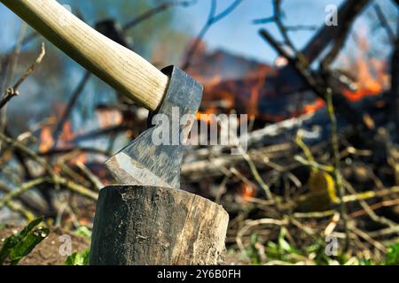 Axt, die im Baumstamm aus nächster Nähe eingebettet ist, brennendes Lagerfeuer im Hintergrund, Outdoor-Aktivitäten wie Holzhacken, Spalten von Stämmen oder Envir Stockfoto