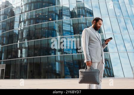 Geschäftsmann in grauem Anzug, der vor einem großen Glasbürogebäude steht, in das Smartphone vertieft, während er eine Lederaktentasche hält Stockfoto