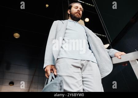 Geschäftsreisender verlassen modernes Gebäude, halten Tasche in der Hand, blicken nach vorne, zeigen Selbstvertrauen und urbanen Lebensstil, in formeller Kleidung gekleidet, Sonnenbrille auf dem Kopf Stockfoto