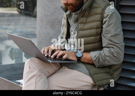 Bärtiger Mann, der draußen auf einer Steinbank sitzt, Hände auf der Laptop-Tastatur, grüne Weste und lässige Kleidung trägt, konzentriert auf den Bildschirm in urbaner Umgebung Stockfoto