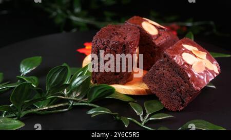 Weiche Schokoladen-Brownie-Stücke mit knusprigen Mandelbelag, serviert auf einem Holztablett mit grünen und roten Blattdekorationen, um ein natürliches Ambiente zu schaffen. Stockfoto