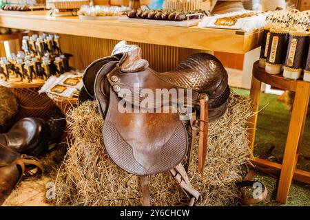 Ein Vintage-Ledersattel auf einem Heuballen in rustikaler Umgebung mit verschiedenen Gegenständen auf einem Holztisch im Hintergrund. Stockfoto