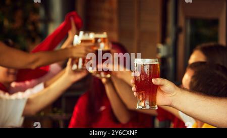 Zwei Gläser klirren, während Sportfans anstoßen. Verschwommene Figuren im Hintergrund verleihen der Szene fröhlichen Jubels Tiefe. Selektiver Fokus. Treffen mit alten Freunden. Stockfoto