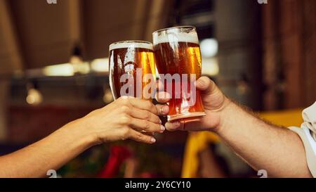 Zwei Hände klirren auf die Brille, Schaum verschüttet. Unscharfe, jubelnde Fans im Hintergrund heben den feierlichen Geist hervor. Treffen mit alten Freunden. Stockfoto