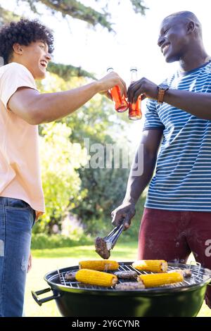 Grillen Sie Mais und Burger, zwei verschiedene männliche Freunde, die mit Getränken beim Grill im Freien toasten Stockfoto