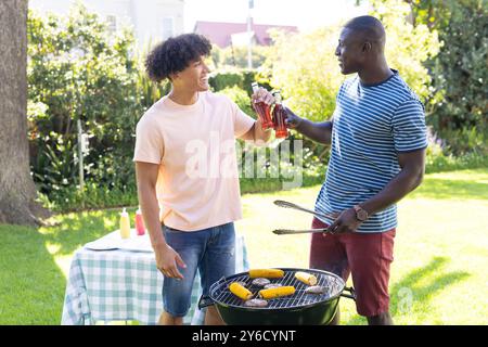 Speisen grillen und mit Getränken toasten, zwei verschiedene männliche Freunde genießen Outdoor Barbecue Stockfoto