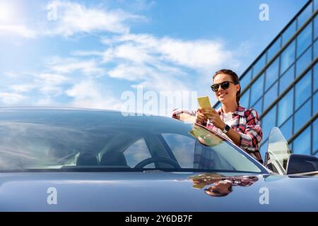 Eine Autofahrerin steht neben ihrem neuen Fahrzeug und nutzt ihr Smartphone vor einem Business Center. Stockfoto