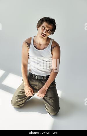 Ein junger Mann mit auffälligen Tätowierungen sitzt in einem Tanktop und taucht in einen nachdenklichen Moment vor einer minimalistischen Kulisse ein. Stockfoto