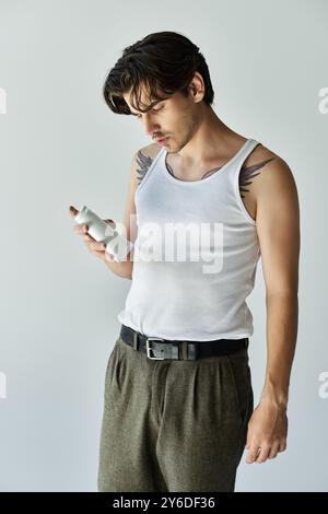 Ein junger, gutaussehender Mann mit einem Tattoo untersucht ein Selbstpflegeprodukt, während er lässig vor grauem Hintergrund gekleidet ist. Stockfoto