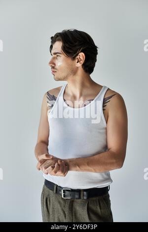 Dieser auffällige junge Mann steht selbstbewusst und zeigt seine Tattoos in einem lässigen Tanktop vor grauem Hintergrund. Stockfoto