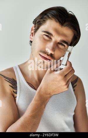 Ein junger Mann mit Tätowierungen trägt ein Tanktop, während er nachdenklich schaut, eingerahmt von einem einfachen grauen Hintergrund. Stockfoto