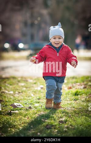 Ein kleiner Junge in einer leuchtend roten Jacke und einem grauen Hut rennt fröhlich durch einen üppig grünen Park, sein Lachen hallt in der frischen Herbstluft, umgeben von B Stockfoto