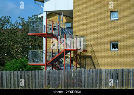 Eine Feuertreppe aus rotem Metall ist an einem Backsteingebäude angebracht und zeigt einen offenen Außenbereich mit Grün und blauem Himmel im Hintergrund Stockfoto