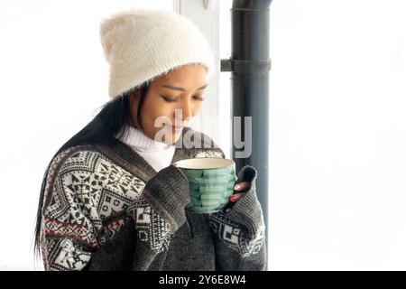 Junge Frau in Winterkleidung, die einen Becher auf der Terrasse hält, frischer Schnee im Hintergrund Stockfoto