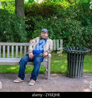 Der Mann entspannt sich auf einer Holzbank in einem grünen Garten und genießt das warme Sonnenlicht, während er neben einem dekorativen Mülleimer sitzt, umgeben von Laub Stockfoto