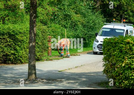 Ein Arbeiter in Sicherheitsausrüstung beugt sich nach unten, um sich in der Nähe eines Hydranten mit einem weißen Fahrzeug auf einem von Bäumen gesäumten Weg in hellem Sonnenlicht zu versorgen Stockfoto