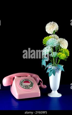 Künstliche Blumen in einer weißen Keramikvase mit einem Vintage-pinkfarbenen Telefon auf schwarzem Hintergrund und blauem Sockel Stockfoto