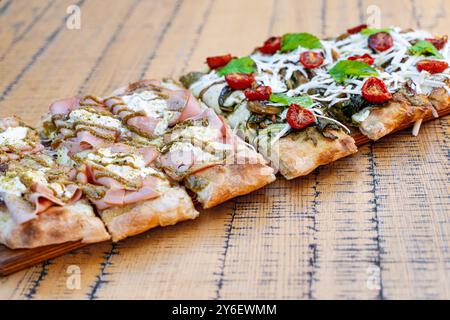 Nahaufnahme von handgefertigten Pizzaschnitten auf einem Holzbrett. Mit Schinken, Kirschtomaten, Kräutern und geschmolzenem Käse. Zeigt eine Vielzahl von Belägen und Texten Stockfoto