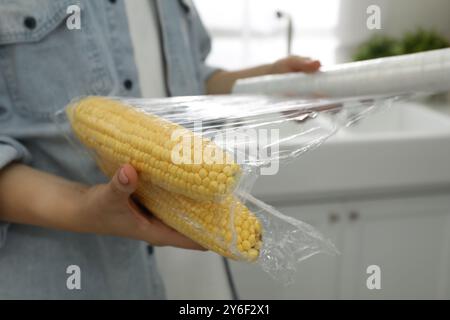 Frau, die Plastikfutterverpackung über Maiskolben in der Küche legt, Nahaufnahme Stockfoto