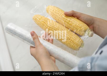 Frau, die Plastikfutterverpackung über Maiskolben in der Küche legt, Nahaufnahme Stockfoto