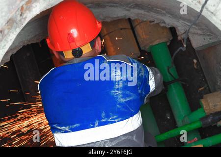 Ein Arbeiter, der einen leuchtend orangefarbenen Helm und Schutzkleidung trägt, ist in einem unterirdischen Tunnel an Reparaturarbeiten beteiligt. Funken fliegen, wenn der Arbeiter Maschinen in der Nähe grüner Rohre bedient, was die Art der Aufgabe unterstreicht. Stockfoto