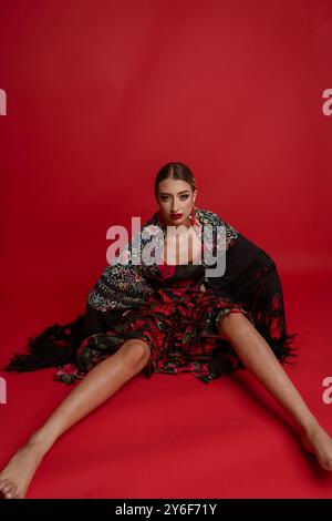 Eine junge Frau in einem traditionellen Blumenkleid und Schal sitzt auf rotem Hintergrund und zeigt Mode und Kultur. Stockfoto