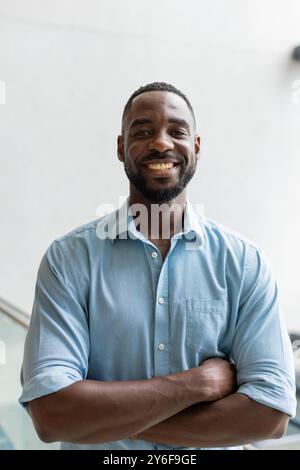 Lächelnder afroamerikaner in blauem Hemd, der sich die Arme kreuzt und selbstbewusst im Büro posiert Stockfoto