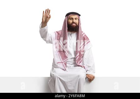 Saudi-arabischer Mann sitzt auf einer leeren Tafel und winkt isoliert auf weißem Hintergrund Stockfoto