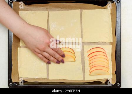 Legen Sie Apfelscheiben von Hand auf Blätterteigquadrate auf ein mit Backpapier ausgelegtes Backblech. Konzept des selbstgemachten Backens und Kochens. Stockfoto