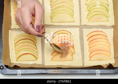 Nahaufnahme einer Hand, die Zucker und Zimt auf geschnittene Äpfel auf Teigquadrate streut, auf ein Backblech gelegt. Konzept des selbstgemachten Backens. Stockfoto