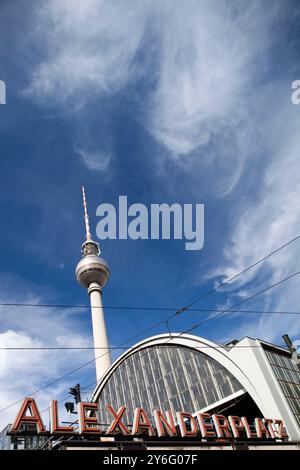 Das Bahnhofsschild Alexanderplatz steht mit dem Fernsehturm, der sich vor einem hellblauen Himmel in Berlin erhebt. Stockfoto