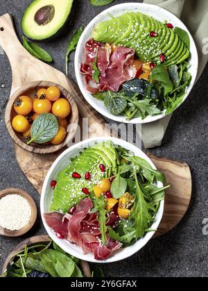 Lebensmittel, Lebensmittel, kohlenstoffarme Schüssel. Frischer Salat mit grünem Spinat, Rucola, Avocado und Schinken serrano in weißer Schüssel, grauer Hintergrund, Blick von oben Stockfoto