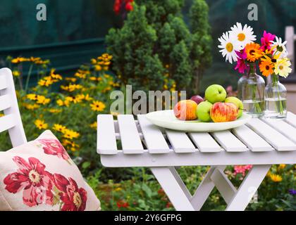 Die Gartenszene verfügt über einen weißen Holztisch mit einem Teller mit Äpfeln, zwei Glasgläser für frische, bunte Gänseblümchen und andere Blumen im Hintergrund, und eine Stockfoto