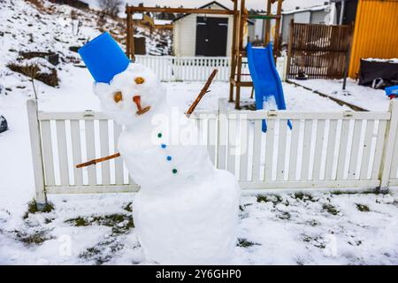 Schneemann mit blauem Eimer auf dem Kopf und Stabarmen, lehnt sich wegen des Abschmelzens bei warmem Wetter, steht im schneebedeckten Garten neben der Kinderrutsche Stockfoto