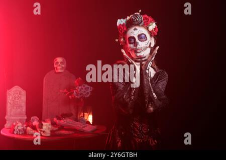 Frau in okkult dekoriertem Atelier während des Day of the Dead Halloween-Events zu Ehren toter Lieben. Eine gruselige Dame, die alte aztekische Gewohnheit macht, Vorfahren zu feiern, mit rotem Licht Hintergrund Stockfoto